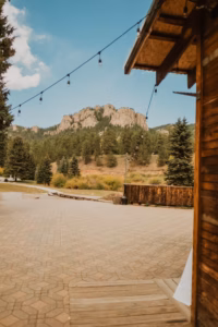 Wooden building with string lights in the foreground, a paved patio, pine trees, and rocky hills under a blue sky in the background.