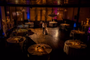 A bride and groom share a first dance in the center of a dimly lit, elegant ballroom with round, candlelit tables surrounding them.