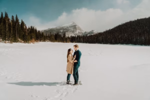 A couple stands facing each other on a snow-covered field surrounded by pine trees and mountains under a cloudy sky.