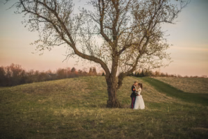 A bride and groom stand together under a large tree in an open grassy field at sunset.
