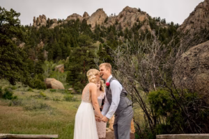 A bride and groom stand outdoors, holding hands and smiling, with rocky mountains and trees in the background.