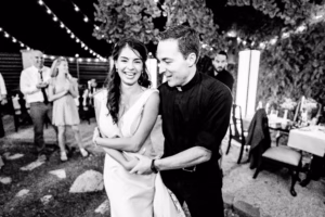 A woman in a white dress and a man in a black shirt with a clerical collar smile and dance together at an outdoor event with string lights and guests in the background.