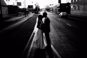 A bride and groom stand facing each other in the middle of an empty street, silhouetted against the bright background.