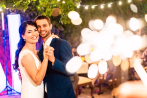 A bride and groom smile and hold hands while dancing at their wedding reception, with warm string lights and blurred decorations in the background.