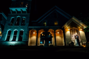 A couple stands embracing at the entrance of a large, illuminated stone building at night, with a fountain visible in the foreground.