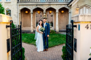 A bride and groom stand together smiling in front of an elegant stone building, holding a bouquet, framed by an open gate.