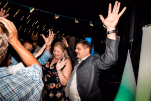 A group of people with raised hands dance and celebrate under string lights at an indoor event.
