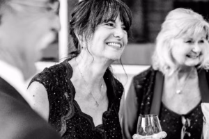 A woman in a dark dress smiles while holding a wine glass, standing next to another woman and a man at a social event.