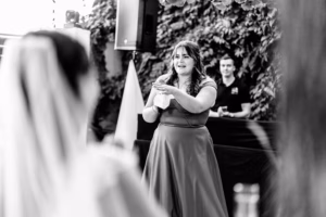 A woman in a formal dress stands and speaks while holding a tissue, addressing a seated bride in the foreground at an event.