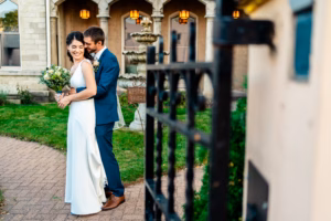 A bride and groom stand together outside a building, smiling as the groom hugs the bride from behind; she holds a bouquet of flowers.