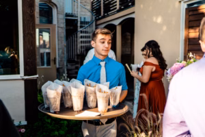 A man in a blue shirt and light tie holds a tray of snack cones at an outdoor gathering, with other people and a building visible in the background.
