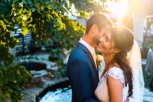 A bride and groom embrace and smile outdoors near a pond, with sunlight streaming through green foliage.