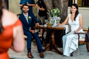 A bride and groom sit at a small table outdoors, listening and smiling, with drinks and flowers on the table during a wedding reception.