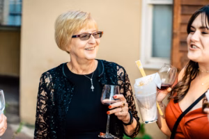 Two women stand outside, holding glasses of red wine and conversing. One woman also holds a container with stirring sticks. Both appear to be at a social gathering.