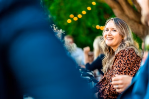 A woman with long blonde hair sits outdoors, smiling, with blurred people in the foreground and string lights and greenery in the background.