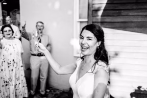 A woman in a white dress holds up a champagne glass and smiles, while people in the background cheer and raise their glasses.
