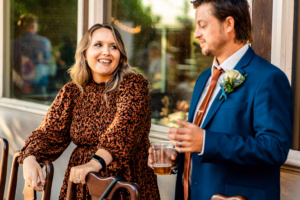 A woman in a patterned dress smiles and talks to a man in a blue suit holding a drink, both standing next to wooden chairs outdoors.