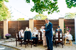 A man in a suit stands and speaks into a microphone at an outdoor dining table surrounded by seated guests during a daytime gathering.