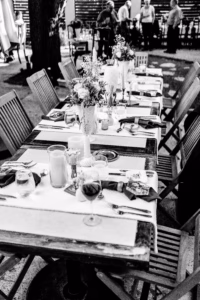 A long dining table set outdoors with flowers, candles, glassware, and napkins, surrounded by wooden chairs; people stand in the background.