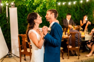 A bride and groom dance closely together outdoors at night, surrounded by guests seated at tables under string lights and greenery.