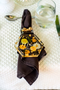 A hexagonal nameplate reading "Mandy Richardson" with yellow flowers rests on a folded dark napkin, next to a glass of water and silverware on a quilted white placemat.
