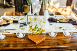 A wooden table set for a meal with glassware, cutlery, candles, and a clear sign featuring yellow and white flowers in the center.