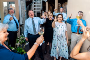 A group of adults standing outdoors raise wine glasses and give thumbs up, appearing to celebrate together in formal attire.