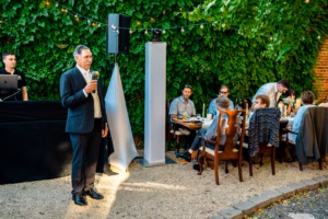 A man in a suit speaks into a microphone outdoors near a table of seated people, with a DJ setup and green leafy background.