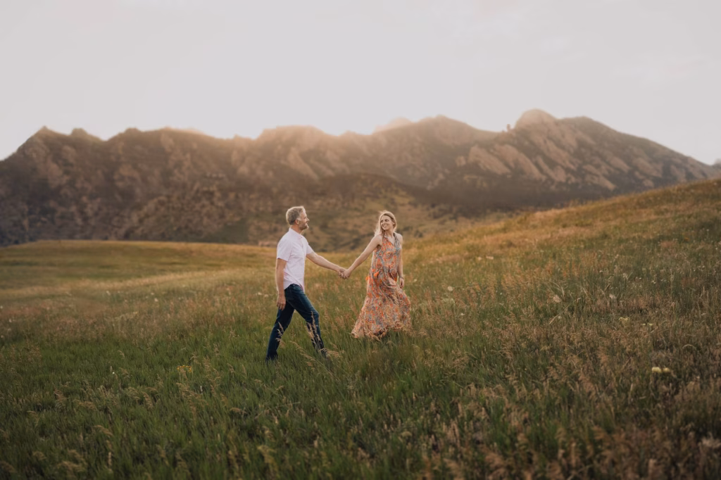 A man and woman holding hands walk through a grassy field with mountains in the background under a cloudy sky.