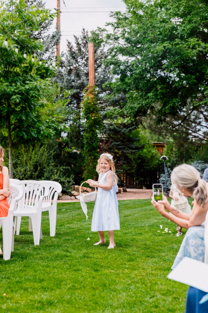A young girl in a white dress walks barefoot on grass, scattering flower petals, while adults watch and take photos at an outdoor event.