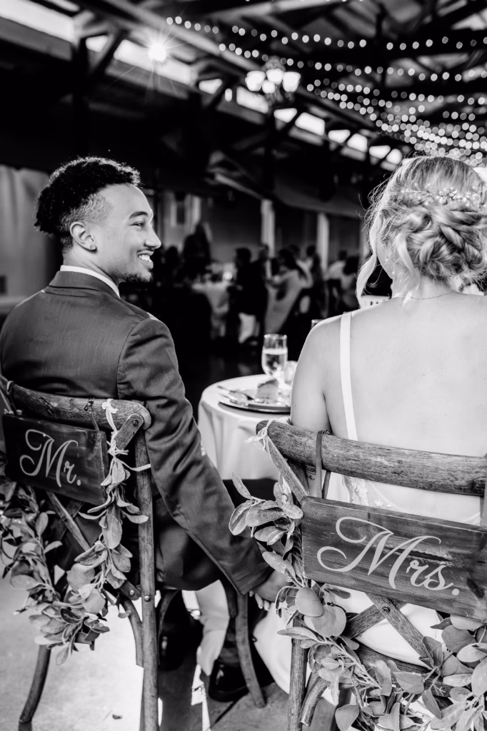 A couple sits at a wedding reception, seen from behind, on decorated chairs labeled "Mr." and "Mrs." with greenery. They face a table with drinks, smiling at each other.