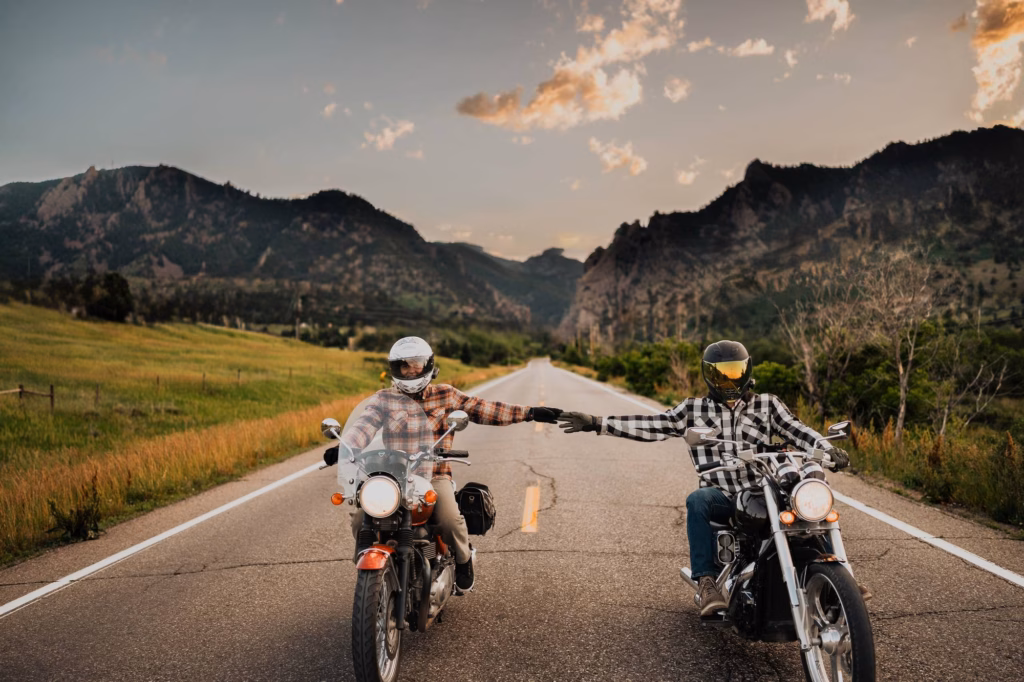 Two people on motorcycles ride side by side on a rural road, extending their arms to touch hands, with mountains and grass fields in the background.