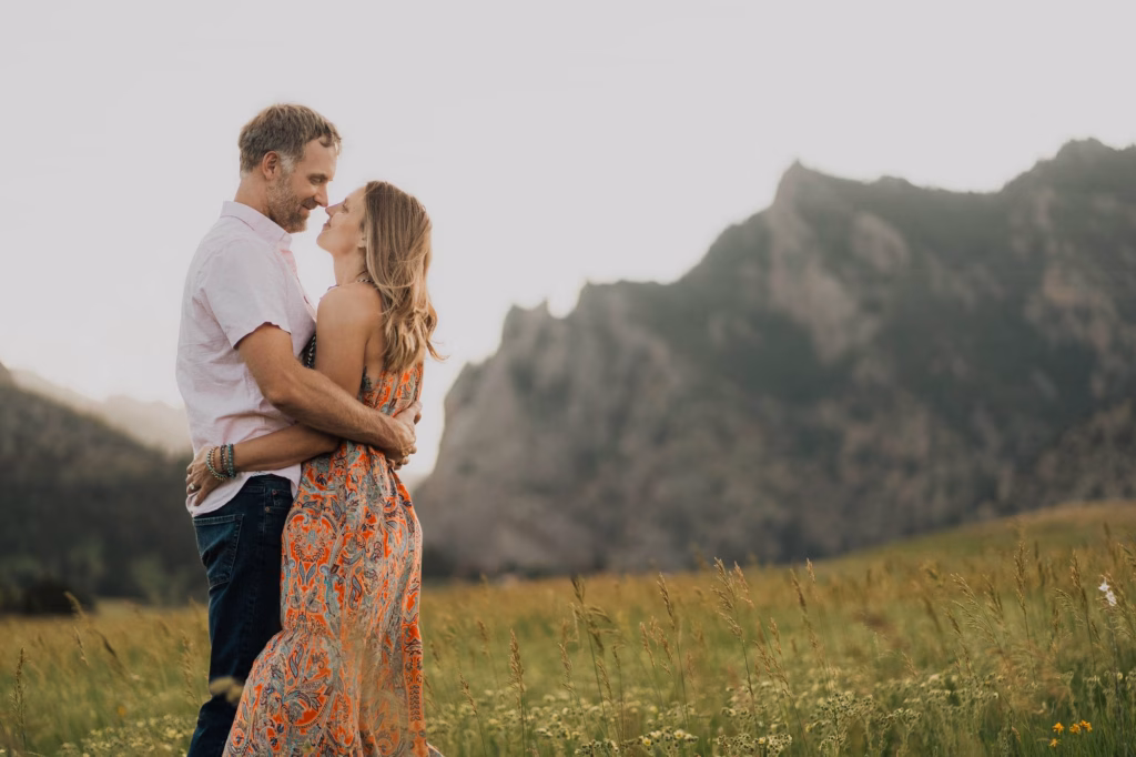 A couple stands embracing in a grassy field with mountains in the background under a clear sky.