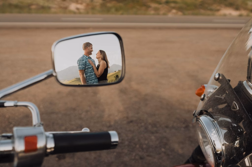 A couple stands facing each other, reflected in a motorcycle mirror, with a road and landscape in the background.