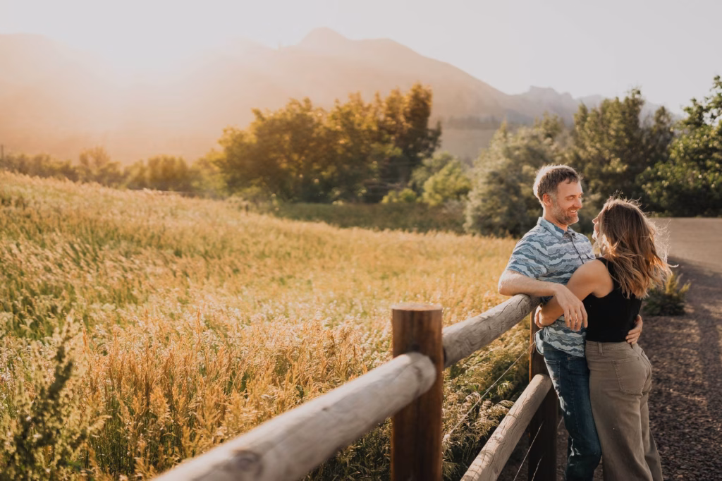 A man and woman stand by a wooden fence in a field at sunset, facing each other and smiling, with mountains and trees in the background.