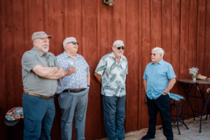 Four older men stand and talk in front of a red wooden wall at the Horstmann Homestead wedding, dressed in casual shirts and sunglasses. A small table with flowers is visible nearby.