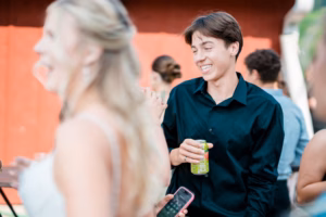 A young man in a black shirt holding a can smiles while talking to others at an outdoor Horstmann Homestead wedding.