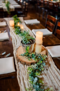A wooden table set for a Horstmann Homestead wedding features white napkins, silverware, a potted succulent on a wood slice, artificial candles, and a greenery runner as the centerpiece.