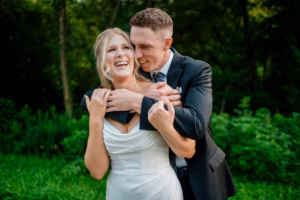 A man in a suit hugs a woman in a white dress from behind as they both smile and laugh, standing outdoors among lush greenery at their Horstmann Homestead wedding.
