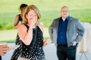 A woman in a black and white dress stands in the foreground, holding her hand to her face at a Horstmann Homestead wedding. Three other people are in the background at an outdoor gathering.