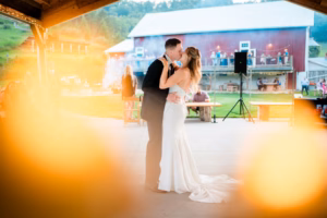 A bride and groom share their first dance under a pavilion at their Horstmann Homestead wedding, with a barn and guests in the background; warm light flares in the foreground.