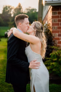 A bride and groom kiss outdoors near a brick building at their Horstmann Homestead wedding, embracing each other in wedding attire with lush greenery in the background.