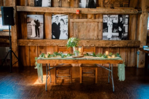 A rustic wooden table with two chairs is decorated with greenery, candles, and flowers, set against a wooden wall adorned with black-and-white Horstmann Homestead wedding photos.