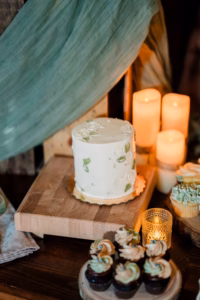 A small white cake with floral decorations sits on a wooden board, surrounded by cupcakes and lit candles on a dark wood table—the perfect centerpiece for a Horstmann Homestead wedding celebration.