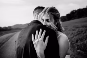 A woman in a wedding dress embraces a man in a suit on a country road at their Horstmann Homestead wedding, proudly showing her engagement ring.