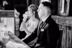 A woman in a white dress wipes her face with a tissue while sitting beside a man in a suit at a wooden table indoors during a Horstmann Homestead wedding.