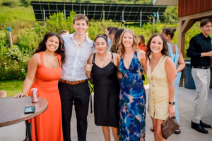 Five people pose and smile for a photo outdoors at a Horstmann Homestead wedding, with a grassy background and other guests visible behind them.