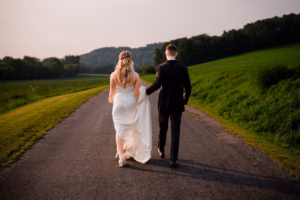 A bride and groom walk hand in hand down a paved country road at their Horstmann Homestead wedding, surrounded by green fields and trees, with their backs facing the camera.