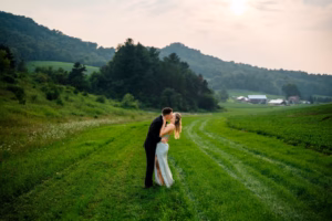 A couple in formal attire kisses on a grassy path at a Horstmann Homestead wedding, surrounded by rolling hills, trees, and farm buildings beneath a bright sky.