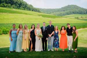 A group of eleven people, including a bride and groom, stand side by side outdoors in front of green hills on a sunny day at a Horstmann Homestead wedding.